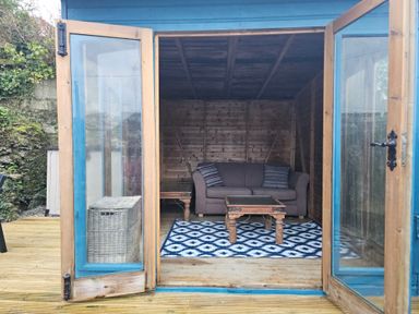 A wooden garden shed with open blue framed double doors showing a sofa with cushions and a wooden coffee table on a patterned rug at Chy An Mor in Mevagissey