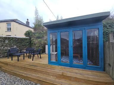 A wooden deck with black outdoor chairs and table next to a blue garden room at Chy An Mor in Mevagissey