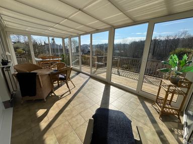 A sunroom with wicker chairs and table a wooden dining table and chairs a plant stand with a plant and large glass windows overlooking a wooden deck at Old Orchard in Saundersfoot