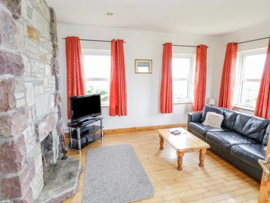 A living room with a stone fireplace a black leather sofa a wooden coffee table a television on a stand and orange curtains at 13 Trawmore Cottages in Keel County Mayo