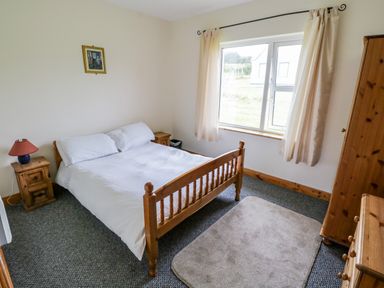 A bedroom with a wooden bed next to a window with curtains and wooden furniture at 13 Trawmore Cottages in Keel County Mayo