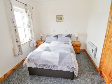 A bedroom with a double bed with patterned bedding two wooden bedside tables with lamps a window with patterned curtains and a small painting on the wall at 13 Trawmore Cottages in Keel County Mayo