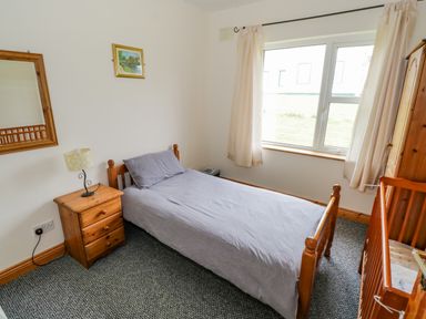 A bedroom with a single bed covered with gray bedding next to a wooden nightstand with a lamp a mirror on the wall and a window with light curtains at 13 Trawmore Cottages Keel County Mayo