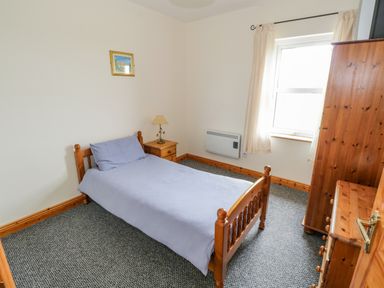 A bedroom with a single wooden bed with a gray blanket and pillow a wooden nightstand with a lamp wooden wardrobe and chest of drawers a window with light curtains and a small framed picture on the wall at 13 Trawmore Cottages Keel County Mayo