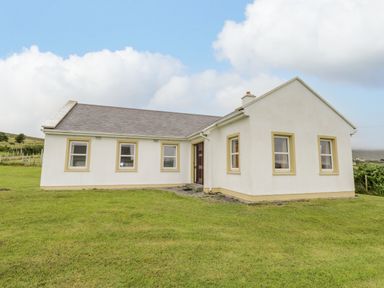 A white single-story house with multiple windows and a brown door on a grassy lawn at 13 Trawmore Cottages in Keel County Mayo