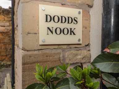 A stone wall with a sign reading Dodds Nook and green plant leaves in the foreground at Dodds Nook in Alnwick