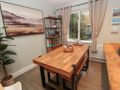A dining area with a wooden table, chairs, shelving unit with books and decor, a window with curtains and a brown sofa at Dodds Nook in Alnwick