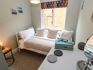 A small bedroom with a bed with white pillows and a beige blanket a window with multicolored blind and a white table with books glasses and coasters at Dodds Nook in Alnwick