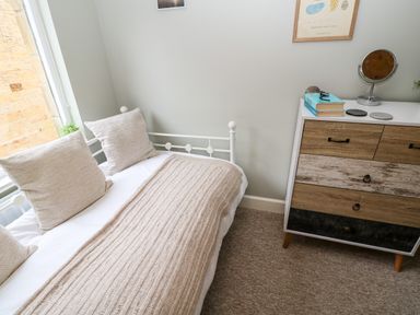 A bedroom with a white metal bed with beige pillows and a beige throw next to a wooden chest of drawers with books and a mirror on top at Dodds Nook in Alnwick