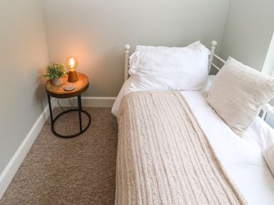A bedroom corner with a bed covered in white and beige linens next to a round wooden side table with a plant and lit lamp at Dodds Nook in Alnwick