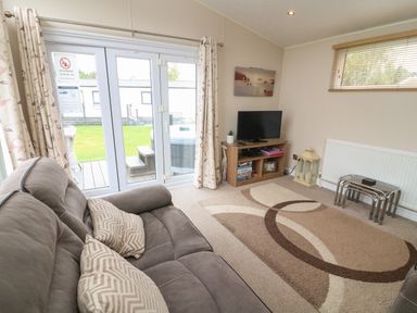A living room with a grey sofa and patterned cushions a TV on a wooden stand a patterned rug and glass patio doors at Waterside Lodge South Lakeland Leisure Village near Carnforth