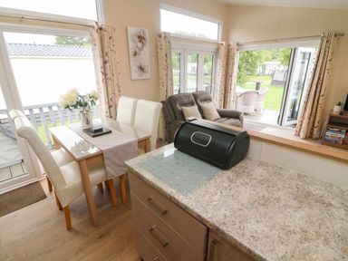 A dining area with a glass table and white chairs next to a living room with a brown sofa and outdoor seating at Waterside Lodge South Lakeland Leisure Village near Carnforth