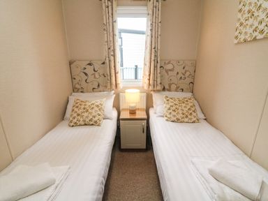 A bedroom with two single beds a nightstand with a lamp and a window with curtains at Waterside Lodge South Lakeland Leisure Village near Carnforth