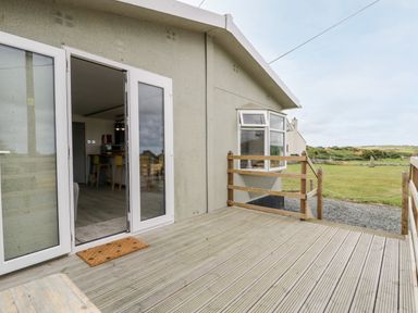 A wooden deck outside a house with open glass doors showing a kitchen with stools at Porthwen Lodge in Bull Bay near Cemaes Bay