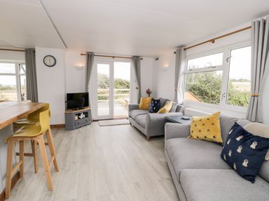 A living room with grey sofas yellow and blue cushions a small TV on a stand and large windows at Porthwen Lodge Bull Bay near Cemaes Bay