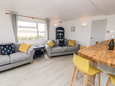A living room with two grey sofas yellow and blue cushions a wooden table with yellow chairs and a window showing fields at Porthwen Lodge in Bull Bay near Cemaes Bay