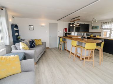 An open living area with a grey sofa, yellow cushions, a wooden kitchen island with yellow stools and black kitchen cabinets at Porthwen Lodge in Bull Bay near Cemaes Bay