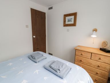 A bedroom with a bed covered by a light blue duvet with white bird prints two folded towels on the bed wooden door wooden dresser with a lamp and a framed picture on the wall at Porthwen Lodge Bull Bay near Cemaes Bay