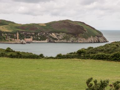 A grassy field overlooking a calm bay with a hillside and old stone buildings at Porthwen Lodge in Bull Bay near Cemaes Bay