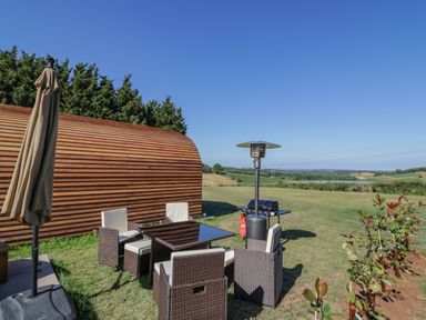 An outdoor seating area with a table chairs an umbrella and a heater near a wooden cabin with a grassy field and hills in the background at Tamina Hillhampton Great Witley