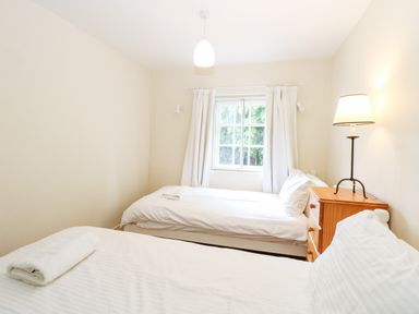 A bedroom with two single beds a wooden bedside table with a lamp and a window with white curtains at Forge House in Cley Next The Sea