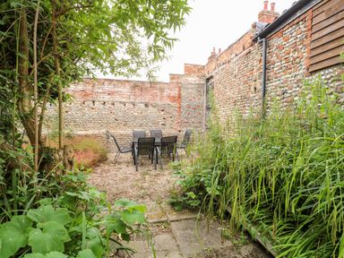 An outdoor patio area with a table and six chairs surrounded by plants and stone walls at Forge House in Cley Next The Sea