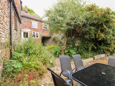 An outdoor garden with a black table and chairs near a brick and flint house surrounded by plants and trees at Forge House in Cley Next The Sea