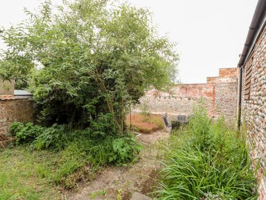 A garden with various green plants a tree in the center and outdoor chairs and table near a stone and brick wall at Forge House in Cley Next The Sea