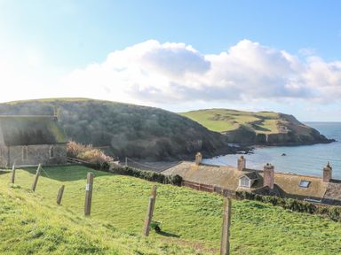A coastal scene with a stone building, grassy hills, rooftops, and the sea at Goody in Hope Cove