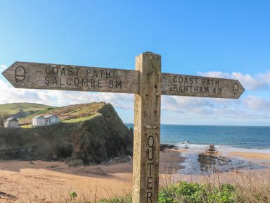 A wooden signpost showing coast path directions to Salcombe and Bantham near a beach with cliffs and a clear sky at Goody in Hope Cove