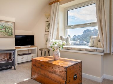 A living room with a wooden chest table with flowers on it a window seat with cushions a television fireplace and framed pictures at Goody in Hope Cove