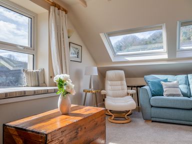A living area with a wooden chest table with a vase of flowers a white armchair a blue sofa with cushions and a window seat with striped cushions at Goody in Hope Cove