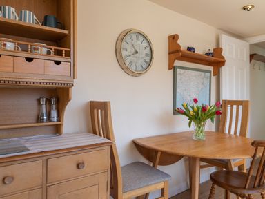 A dining area with a wooden table, chairs, a vase of tulips, wall clock, map, and wooden shelving at Goody in Hope Cove