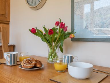 A wooden dining table with tulips in a glass vase cups of juice plates with pastries and a white bowl at Goody in Hope Cove