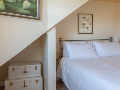 A bedroom with a white bed and pillows two storage chests under stairs and paintings on the walls at Goody in Hope Cove