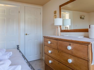 A bedroom with a wooden dresser with a lamp and mirror next to white doors and a bed with folded towels at Goody in Hope Cove