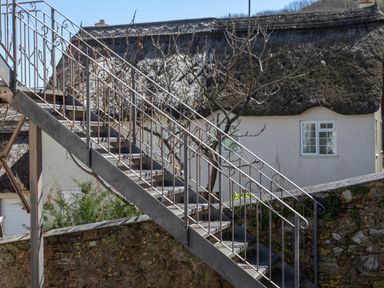 An outdoor metal staircase next to a stone wall with a thatched roof cottage and windows in the background at Goody in Hope Cove
