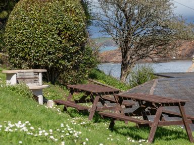 A garden area with wooden picnic tables and a stone barbecue near water at Goody in Hope Cove