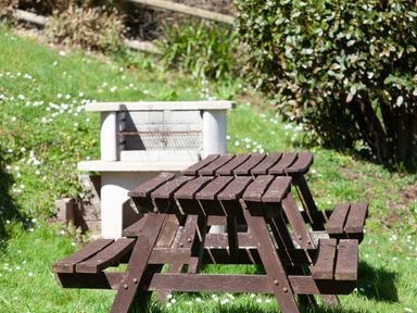 A wooden picnic table with attached benches near a concrete barbecue grill in a grassy garden