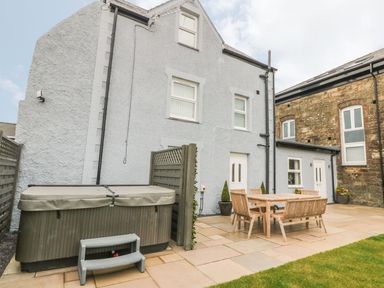 An outdoor patio with a hot tub and wooden dining table with chairs outside a gray house at Fishermans Cottage Malltraeth