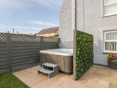 An outdoor hot tub with steps on a patio next to a house with a gray fence and a green privacy screen at Fishermans Cottage in Malltraeth