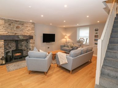 A living room with grey sofas a wooden coffee table a stone fireplace a wall-mounted TV and a staircase at Fishermans Cottage in Malltraeth