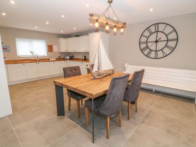 A kitchen and dining area with a wooden table and four chairs a white bench and a large wall clock at Fishermans Cottage in Malltraeth