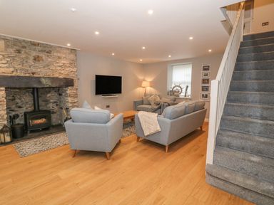 A living room with grey sofas and a wood burning stove next to a staircase at Fishermans Cottage in Malltraeth