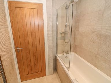 A bathroom with a wooden door a bathtub with a glass shower screen and tiled walls at Fishermans Cottage in Malltraeth