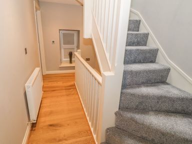 A hallway with wooden flooring a white radiator a window with a vase and carpeted stairs with white railing at Fishermans Cottage in Malltraeth