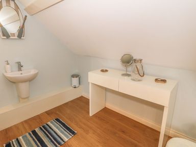 A room with a small sink mirror a white table with a round mirror and glass jar on it a striped rug on a wooden floor at Fishermans Cottage in Malltraeth