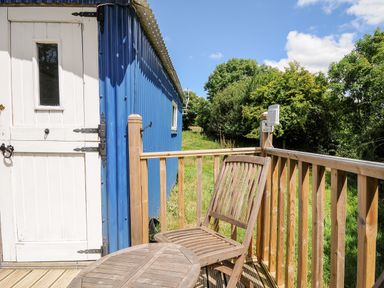 A wooden chair and table on a wooden deck outside a blue building with a white door at Shepherds Hut in Boconnoc near Lostwithiel
