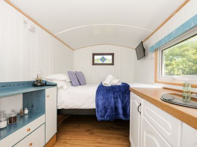 A small room with a bed, pillows, a blue blanket, a cabinet with jars, a sink, and a window at Shepherds Hut Boconnoc near Lostwithiel