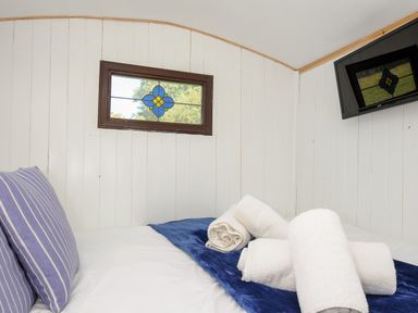 A bed with striped pillows and rolled towels in a white wooden room with a stained glass window and wall mounted TV at Shepherds Hut in Boconnoc near Lostwithiel